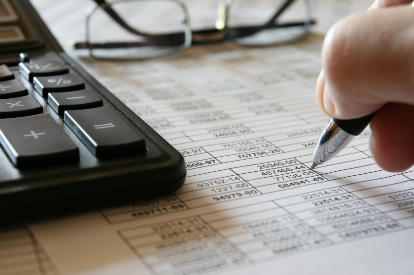 A hand holding a pen over a spreadsheet next to glasses and a keyboard